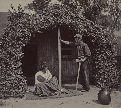 WALTER BURKE, SYDNEY Portrait of Waaka Tamaira, Tokaanu, holding a kakauroa and a Māori woman holding a carved waihaka, at entrance to ivy-covered reed whare