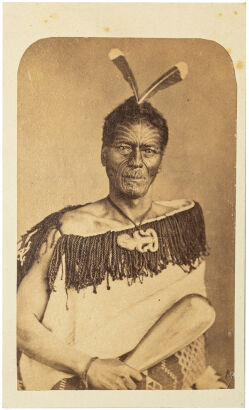 FOY BROS, THAMES Portrait of Māori man with moko, two Huia tail feathers, wearing a Kaitaka aronui with taniko border, heitiki and holding a patu