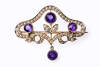 An Edwardian amethyst and pearl brooch, three round amethyst surrounded with half seed pearls set in a floral scroll with a amethyst and seed pearl drop. 15ct yellow gold. Width 40mm. Circa 1910.