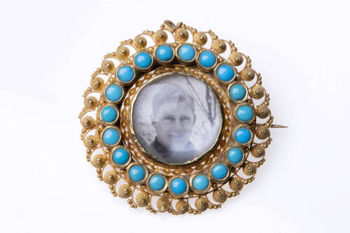 A Victorian turquoise and gold memorium brooch, the target brooch shape with central glassed locket window is surrounded by two borders one with rubover turquoise the outer raised canetille motifs. Diameter 34mm. Circa 1840.