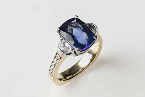 A tanzanite and diamond ring, the cushion cut tanzanite 11mm x 8.2mm of estimated weight 4.25 carats flanked by two half moon shape diamonds and 10 round brilliant diamonds channel set down the band. 18ct white and yellow gold. Weight 8.2 grams. Size M.&nbsp;