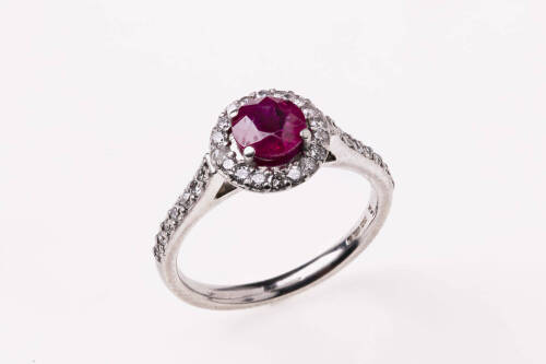 A ruby and diamond ring, the round ruby four claw set above a border of round brilliant diamonds with diamonds down shoulders. 18ct white gold. Weight 5.42 grams. Size M.&nbsp;