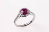 A ruby and diamond ring, the round ruby four claw set above a border of round brilliant diamonds with diamonds down shoulders. 18ct white gold. Weight 5.42 grams. Size M.&nbsp;